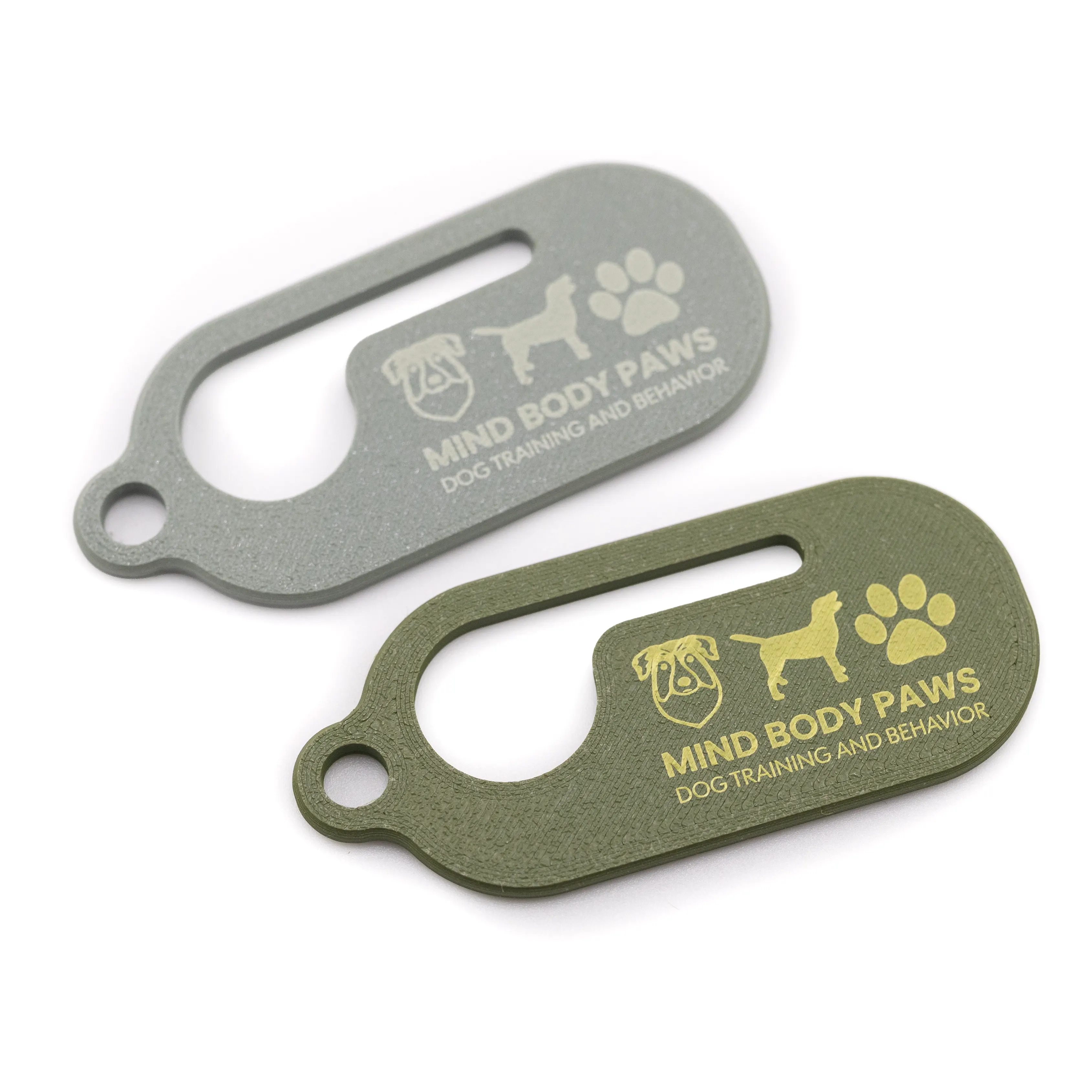 Two 3D-printed dog waste bag holders in sage and olive green, each engraved with the Mind Body Paws logo.